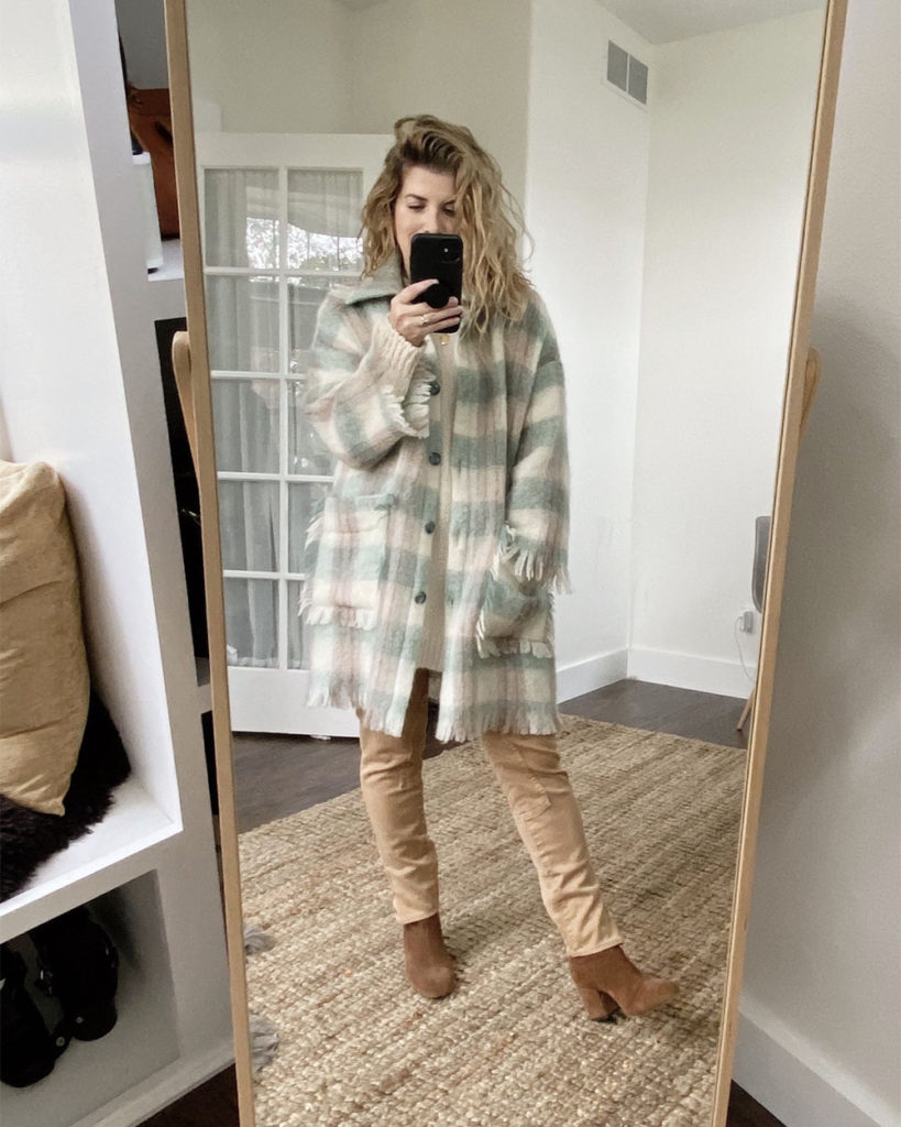 Sweater Outfit 3 - a mirror selfie of a white woman with wavy blonde hair wearing a cream crew neck long sweater over a pair of tan pants. She has a grey scarf tied around he neck and is wearing brown suede boots. She has added a long checked wool coat that has green, cream and tan colors.
