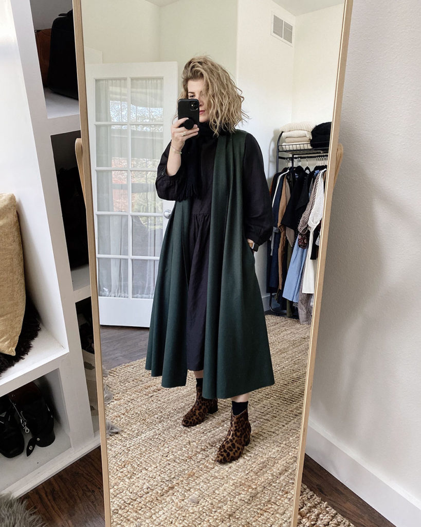 A mirror selfie of a white woman with blond wavy hair who is wearing a long green vest over a long sleeved black dress that is slightly oversized. The dress is midi length and she is wearing leopard print boots.
