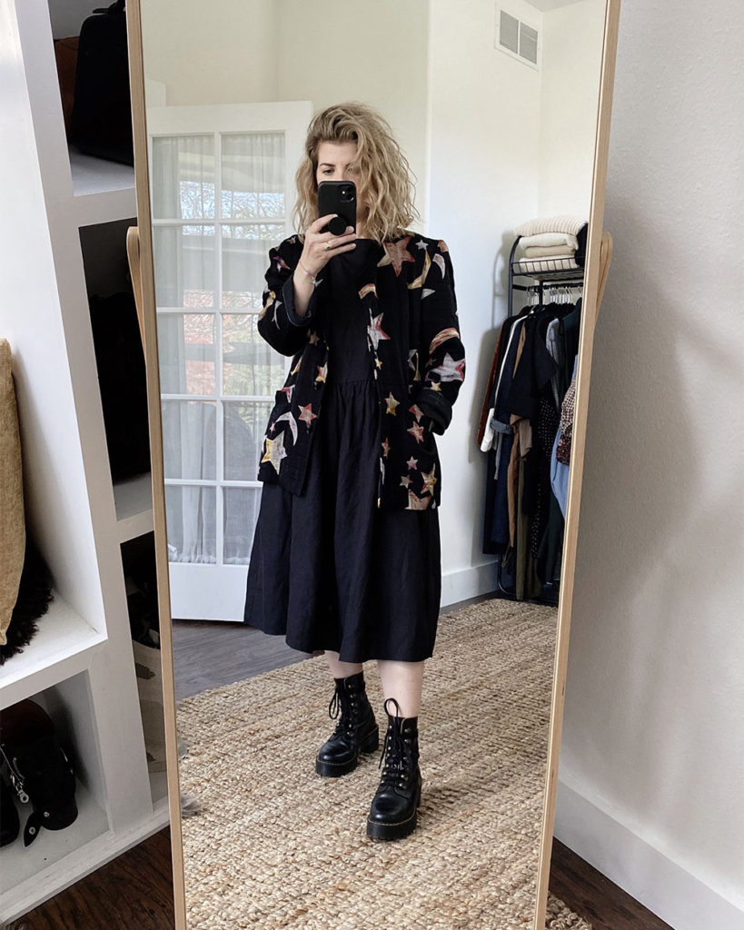 A mirror selfie of a white woman with blond wavy hair who is wearing a black jacket with moons and stars all over it with a black dress that is slightly oversized. The dress is midi length and she is wearing black combat boots.