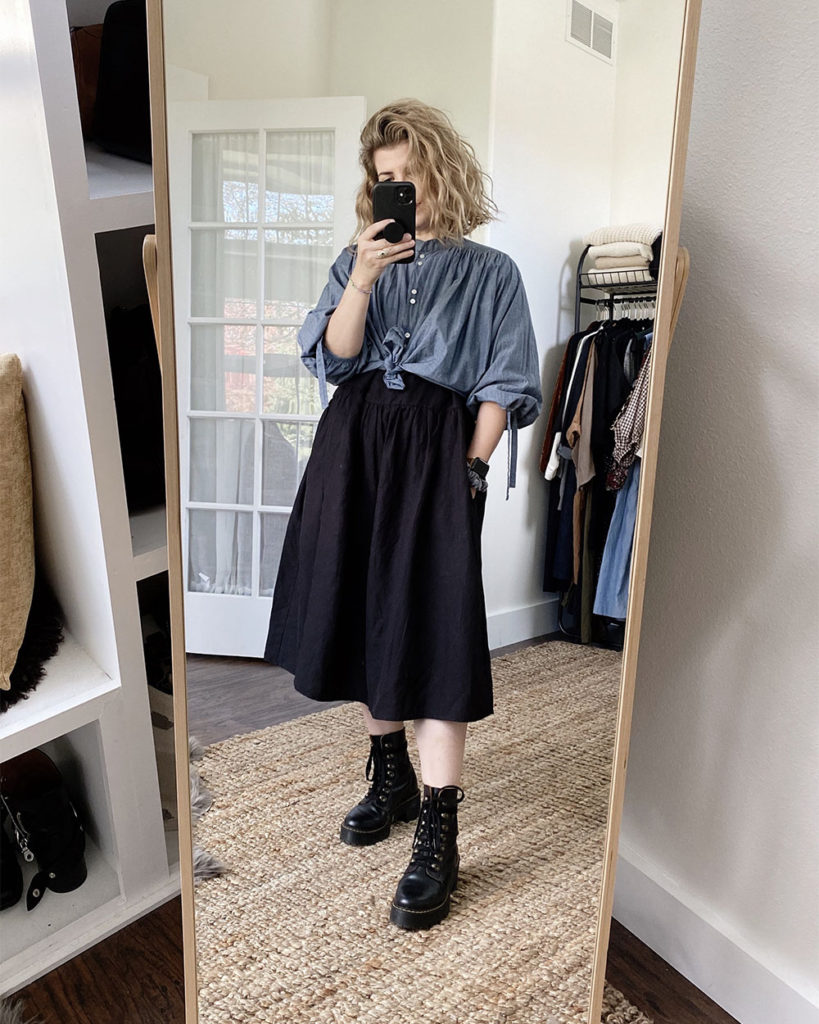 A mirror selfie of a white woman with blond wavy hair who is wearing a blue chambray shirt knotted at the waist over a long sleeved black dress that is slightly oversized. The dress is midi length and she is wearing black combat boots.