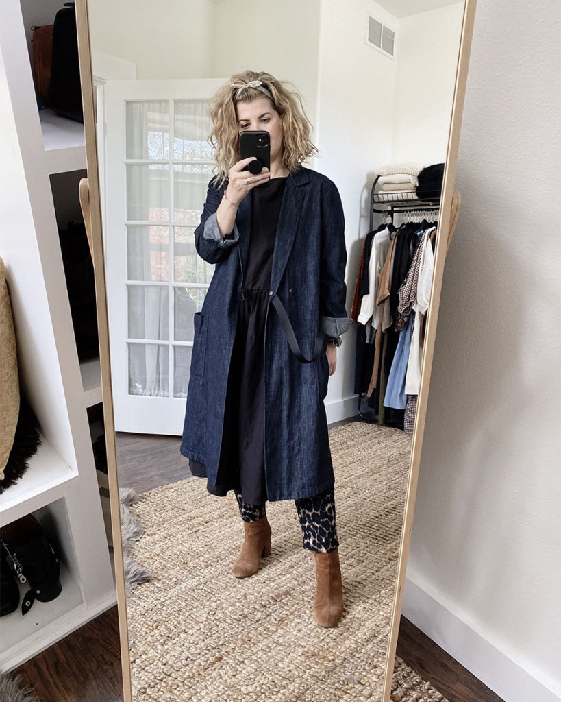 A mirror selfie of a white woman with blond wavy hair who is wearing a long denim jacket over a midi length black dress that is slightly oversized. She is wearing leopard print pants under the dress and brown suede boots.