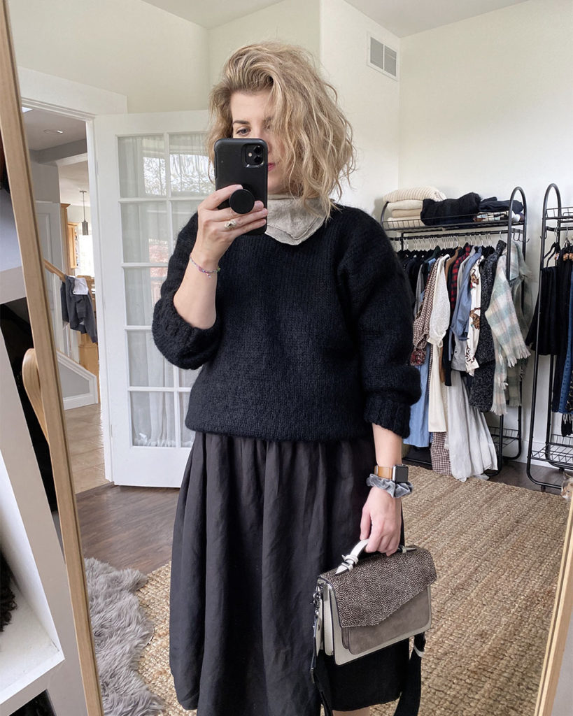 A mirror selfie of a white woman with blond wavy hair who is wearing a black wool sweater over a black dress that is slightly oversized. She has a grey bandana around her neck and is holding a grey satchel handbag. you cannot see the bottom part of the outfit.