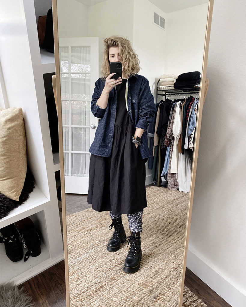 A mirror selfie of a white woman with blond wavy hair who is wearing an oversized navy flannel shirt over a  long sleeved black dress that is slightly oversized. She is wearing black and white printed pants under the dress with black combat boots.