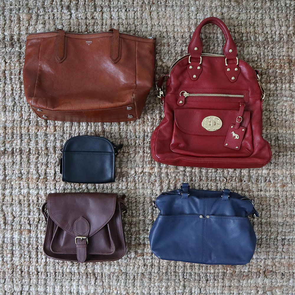 The handbags that I am discarding laid out on a tan rug. There are 2 brown ones, a small black one, a red one and a blue one.