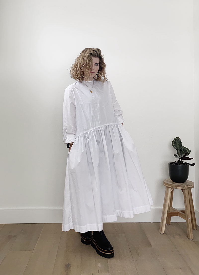 A white woman with dark blond wavy hair is standing in front of a white wall and wearing a long white dress with a dropped pleated waist and long sleeves with black platform boots. She has both of her hands in her pockets.