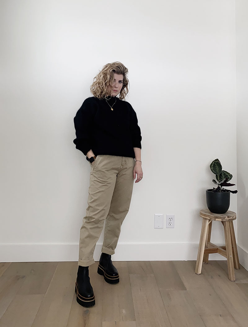 A white woman with dark blond wavy hair is standing in front of a white wall and wearing a black crew neck sweater with beige pants and black platform boots. She is slight turned away from the camera and her right hand is in her pocket. The pants and boots were the first items I bought in my month long shopping spree that led to my resolve to embrace a slower wardrobe.