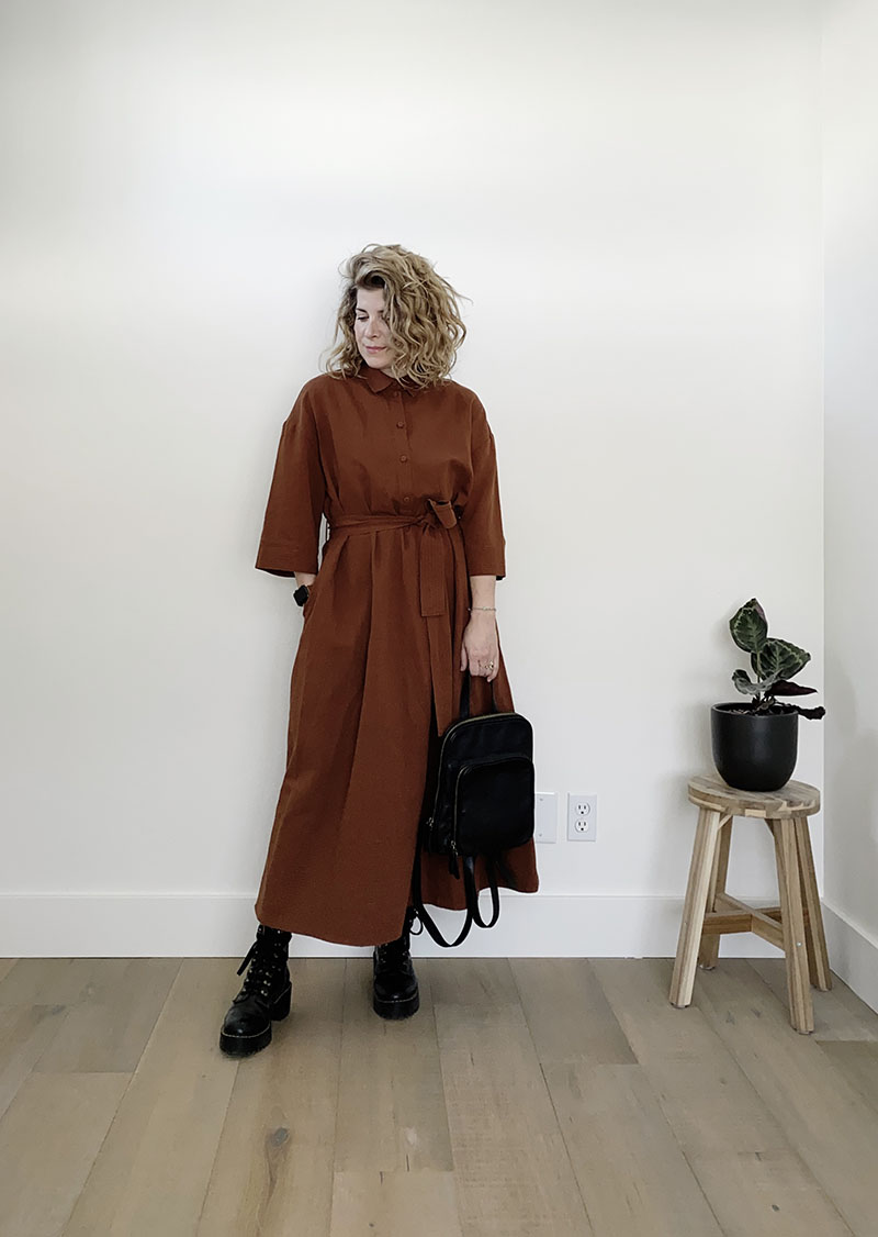 A white woman with dark blond wavy hair is standing in front of a white wall and wearing a long brown dress with a belted waist and three quarter length sleeves with black platform lace up boots. She has one hand in her pocket and she is holding a black backpack with the other hand.