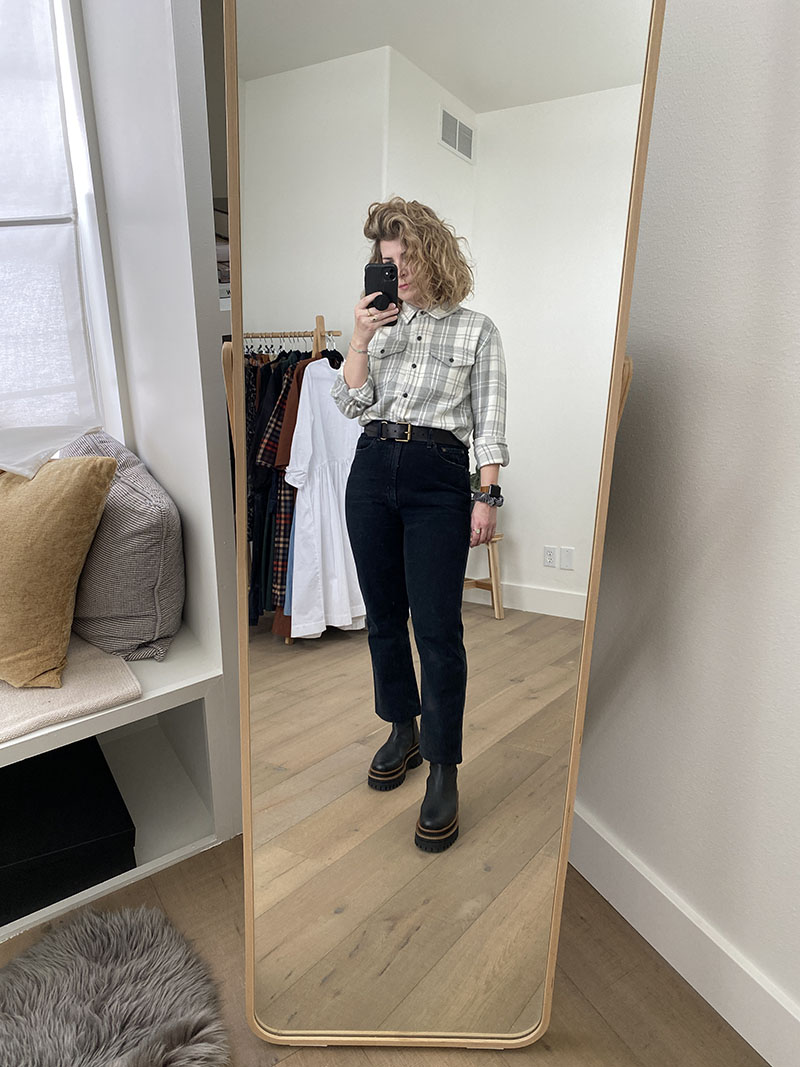 A mirror selfie of a white woman with dark blond wavy hair who is wearing a grey and white checkered shirt with black jeans, a black belt and black platform boots. 