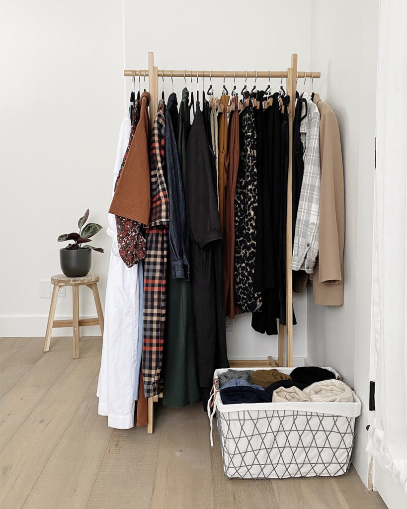 A wooden clothing rack that holds a the majority of my capsule wardrobe items including dresses, trousers, and a few other items such as a shirt, long vest and blazer. There is a basket of sweaters that are rolled up at the base of the clothing rack.