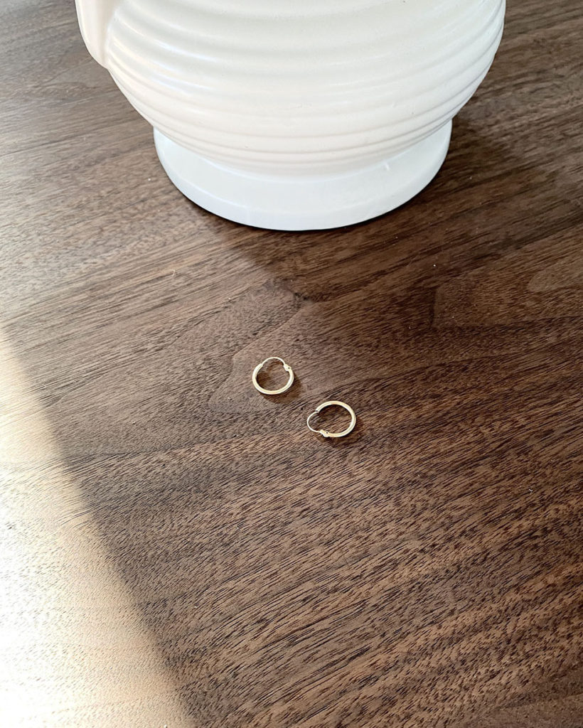 A pair of small gold hoops is laid at the base of a white vase and on top of a dark brown walnut table. You can partially see the vase and table.