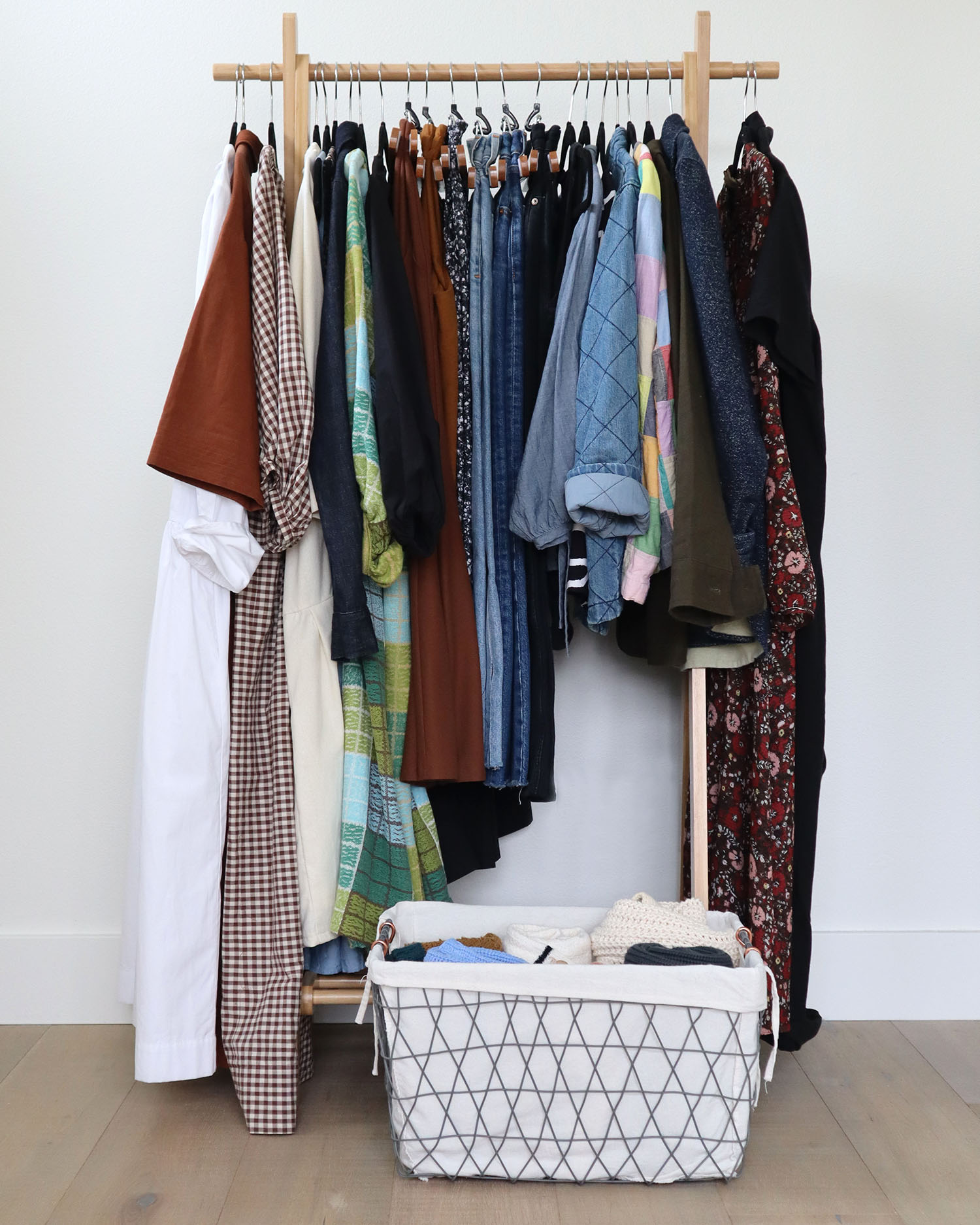 A wooden clothing rack holding 24 items of clothing is in front of a light wall. There is a wire basket in front of it, off to the right, which holds 6 sweaters.