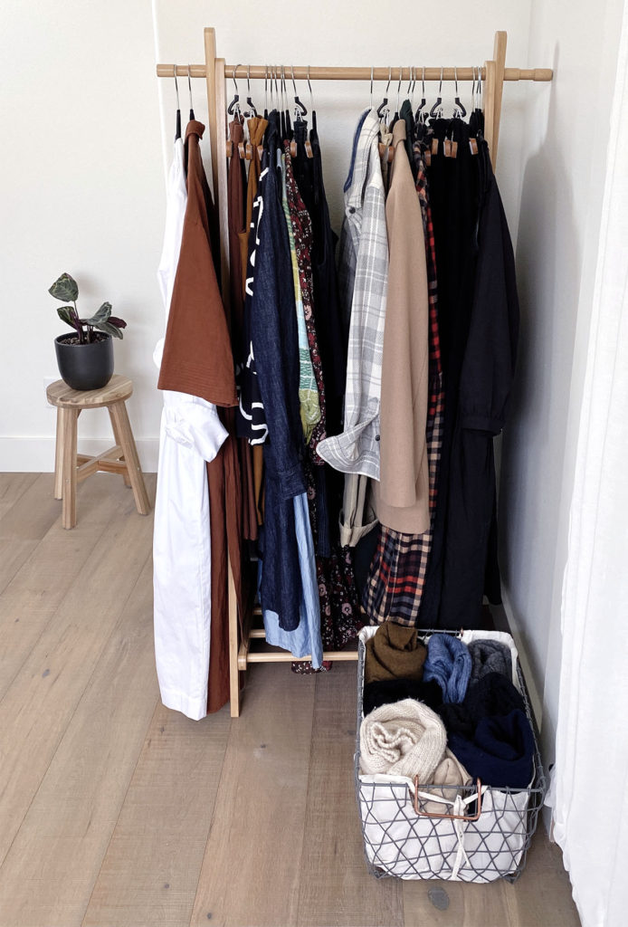 A wooden rack contains clothing from my winter seasonal wardrobe that have been divided into 2 sections: one section to stay and one to be put away. There is a basket of sweaters at the base of the rack and a prayer plant of a stool off to the left.
