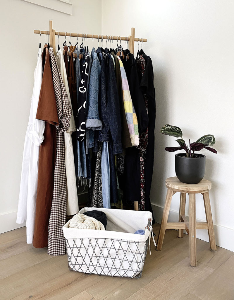 A wooden clothing rack contains clothing from my spring seasonal wardrobe. There is a basket of sweaters at the base of the rack and a prayer plant of a stool off to the right.