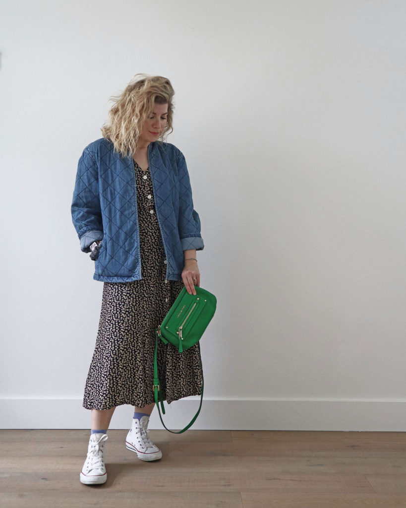 A white woman with blond wavy hair is standing in front of a light wall and wearing a blue quilted denim jacket over a navy button front dress with small tan flowers from her spring capsule wardrobe. She is wearing white converse sneakers with bright blue socks and is holding a bright green handbag.