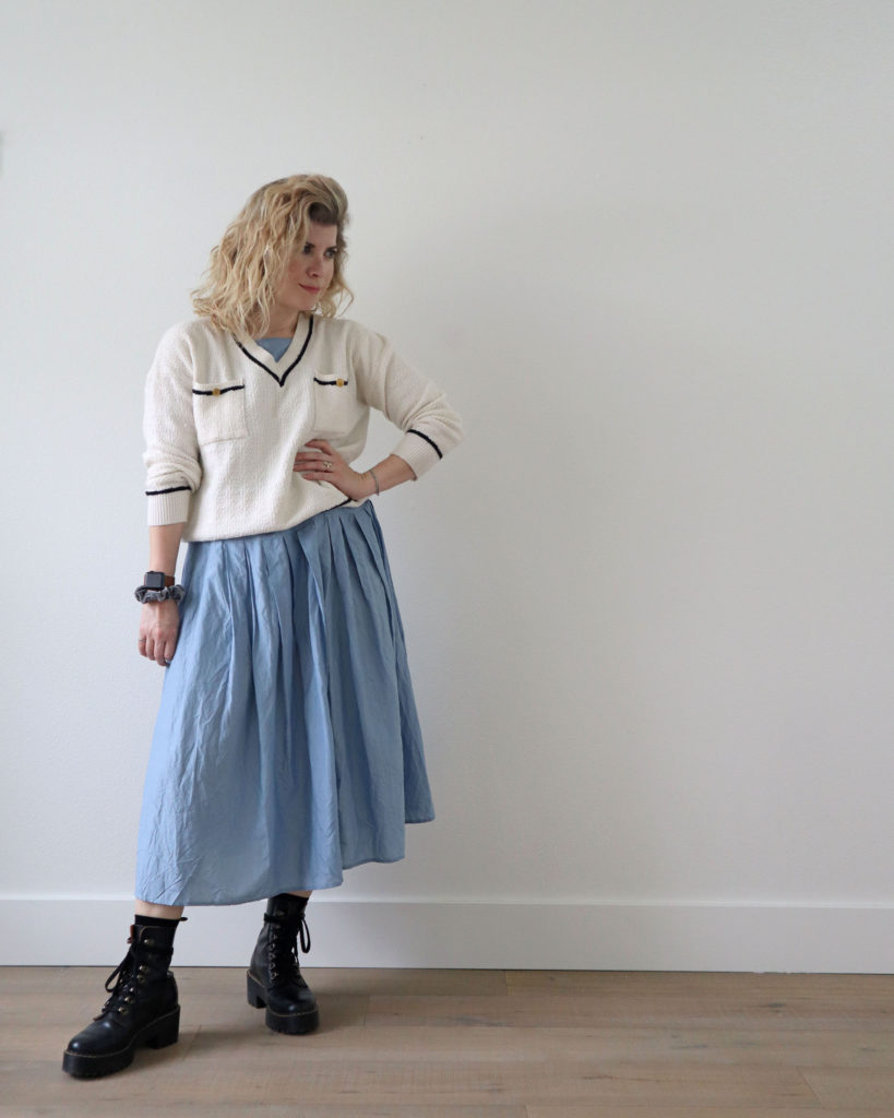 A white woman with blond wavy hair is standing in front of a light wall and wearing a white v neck sweater over a light blue long dress from her spring capsule. She is wearing black combat boots. There is a navy blue border around the waist, sleeves and neck of the sweater and gold buttons on the front pockets.