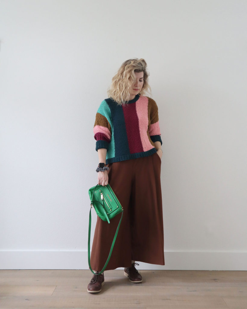 A white woman with blond wavy hair is standing in front of a light wall and wearing a colorful sweater with stripes of brown, pink, Burgundy, teal and mint green with a pair of brown pleated, wide legged pants and brown Oxford shoes. She is holding a bright green purse. This is the fourth outfit from her spring capsule.