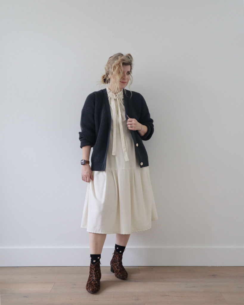 A white woman with her blond wavy hair pulled back into a low ponytail is standing in front of a light wall and wearing a grey cardigan with cream buttons over a cream colored midi length pleated dress from her spring capsule. She is wearing black socks with little pink tufts and brown and black leopard print boots.
