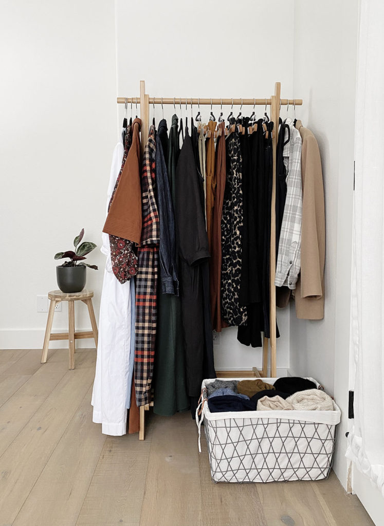 A wooden clothing rack that holds my winter capsule wardrobe. There is a wire basket at the base of the rack with sweaters in it.