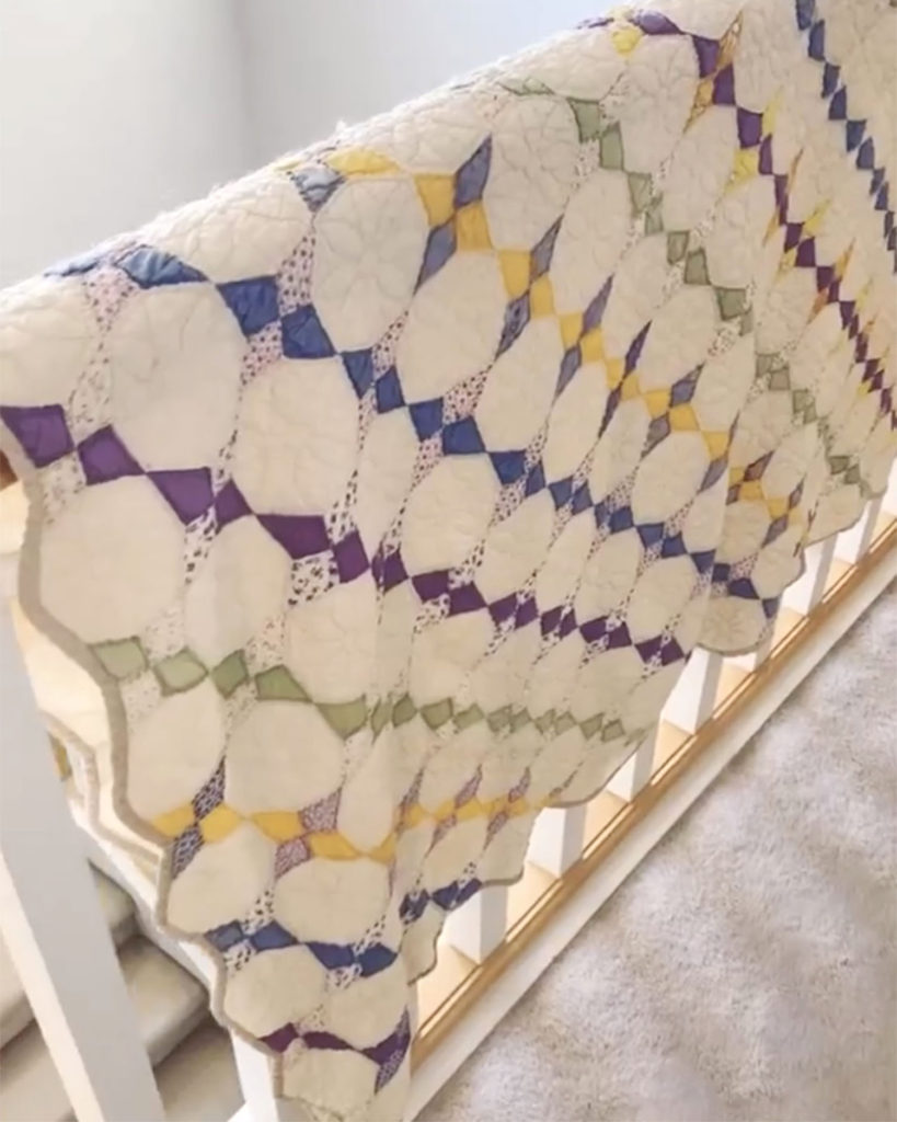 A mostly white vintage quilt hanging over a stair rail. There are various diamond shapes of purple, blue, green and yellow all over it. 