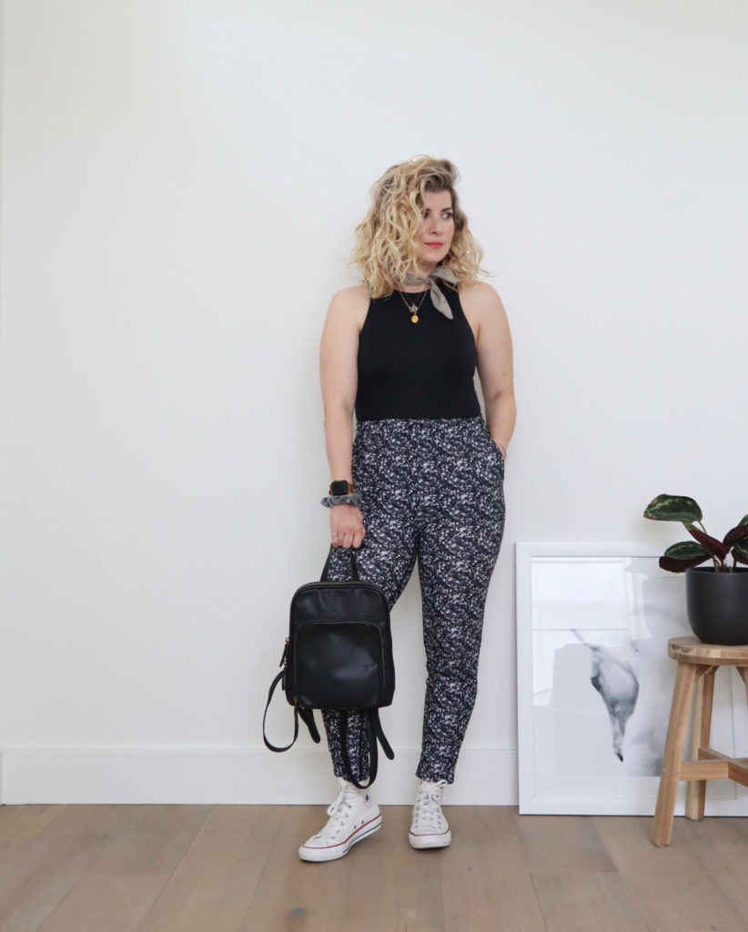 How to style converse outfit 1 - A white woman with blond wavy hair is standing in front of a white wall and wearing a black bodysuit with black and white patterned pants. She is holding a black backpack in her right hand and wearing white high top sneakers. There is a partial picture of a horse and a potted plant on a wooden stool off to her left.