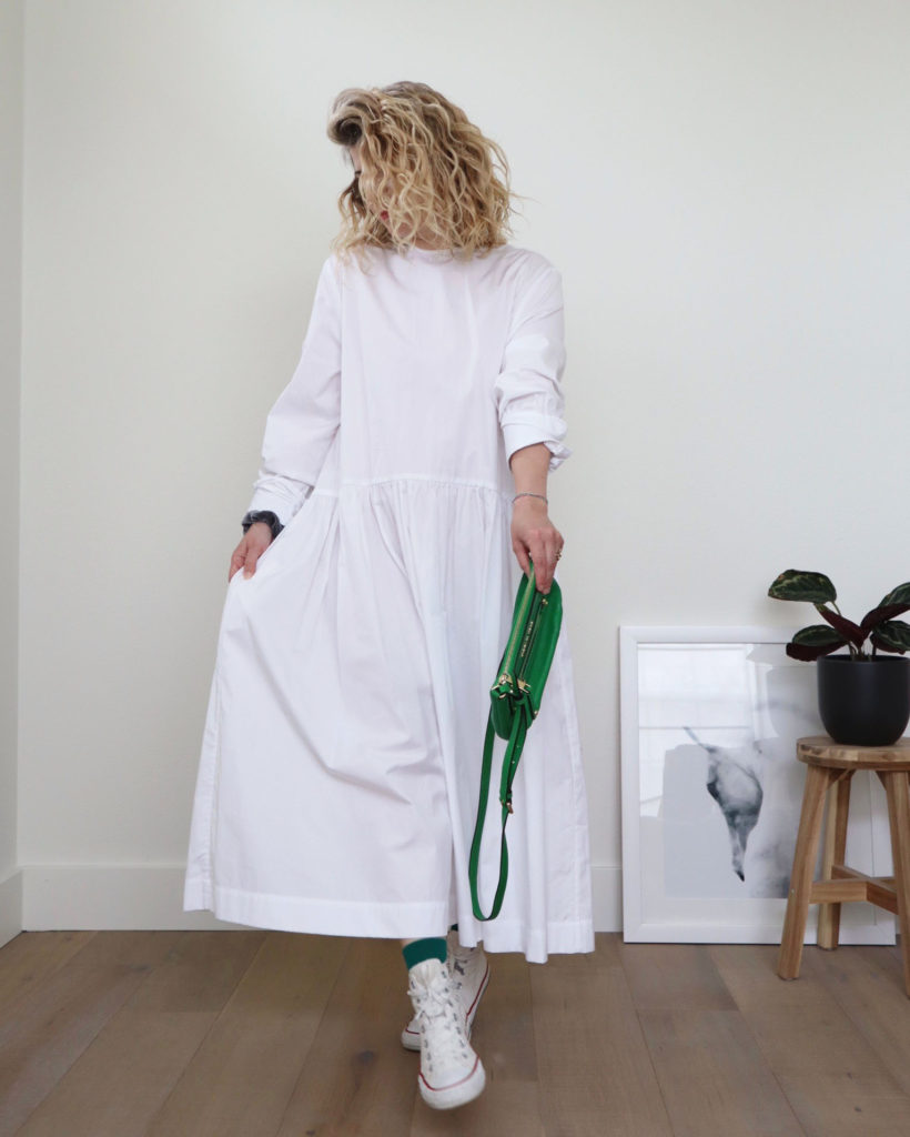 How to style converse outfit 2 - A white woman with blond wavy hair is standing in front of a white wall and wearing a long white dress with a dropped waist and long sleeves. she is holding a green handbag in her left hand and is wearing green socks and white canvas high tops. There is a partial picture of a horse and a potted plant on a wooden stool off to her left.