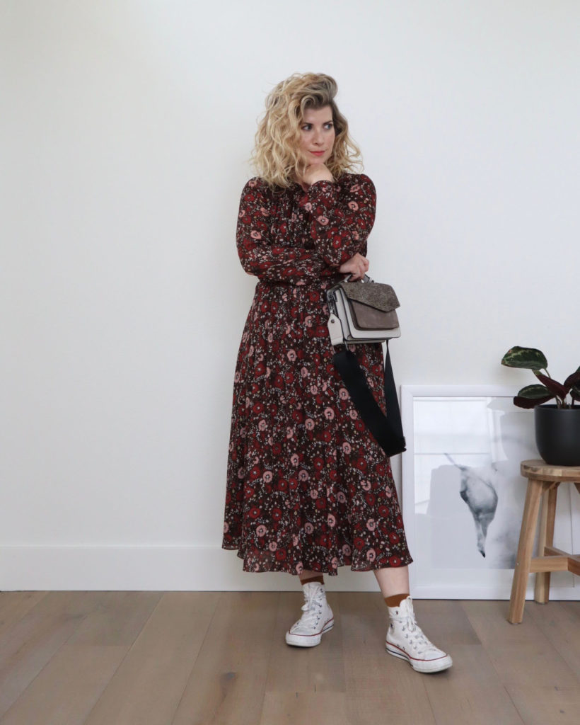 How to style converse outfit 3 - A white woman with blond wavy hair is standing in front of a white wall and wearing a long floral dress with colors of brown, pink and red. she has one arm across her body and it is holding a grey handbag while the elbow of her other arm rests on it and is under her chin. She is wearing brown socks and white high tops. There is a partial picture of a horse and a potted plant on a wooden stool off to her left.