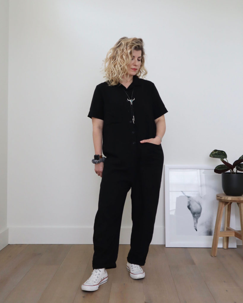How to style converse outfit 4 - A white woman with blond wavy hair is standing in front of a white wall and wearing a a black short sleeved jumpsuit with a silver bull head bobo tie and white high tops. Her left hand is in her pocket. There is a partial picture of a horse and a potted plant on a wooden stool off to her left.