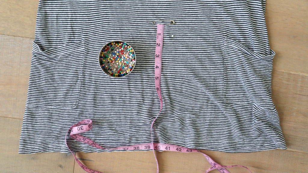 The first step to crop a top is to determine where to cut. This image is of the bottom portion of the tunic that is laid out on a wood floor shot from above. There is a safety pin at the crop limit, a pink measuring tape and a container of colorful sewing pins.