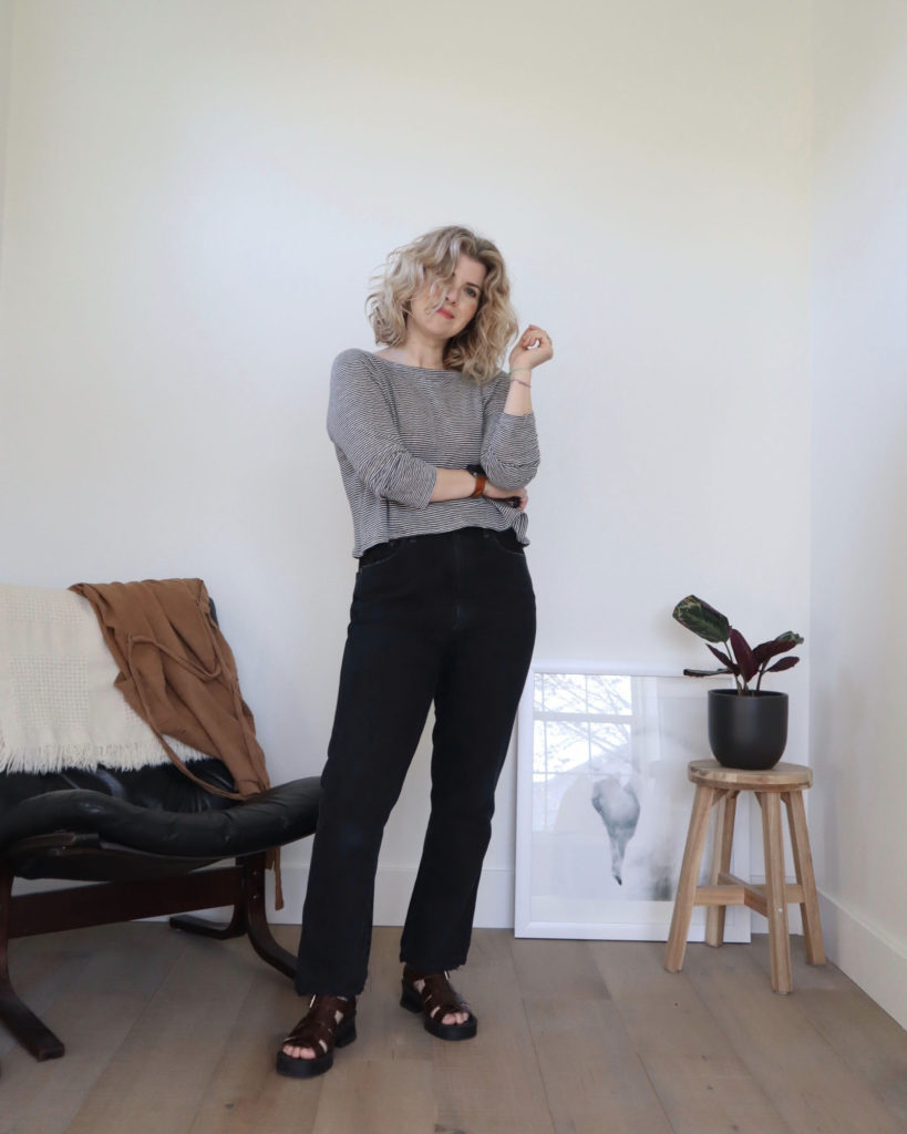 A white woman with short blonde wavy hair is looking at the camera. She is wearing a long sleeved black and white cropped top with black jeans with brown strappy platform sandals. There is a black chair behind her to the left and a prayer plant on a wooden stool behind her to the right. This is the final result of her tutorial on how to crop a top.