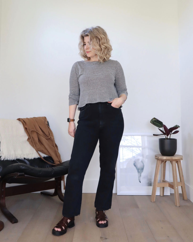 A white woman with short blonde wavy hair is looking off to her right (the photo's left). She is wearing a long sleeved black and white cropped top with black jeans with brown strappy platform sandals. There is a black chair behind her to the left and a prayer plant on a wooden stool behind her to the right. She is using this top as a tutorial on how to crop a top.