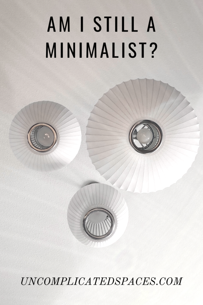 A photo of 3 round white light shades hanging from a ceiling of varying sizes taken from below. Rays of sunlight stream across them. Above the lights is the text "am I still a minimalist?"