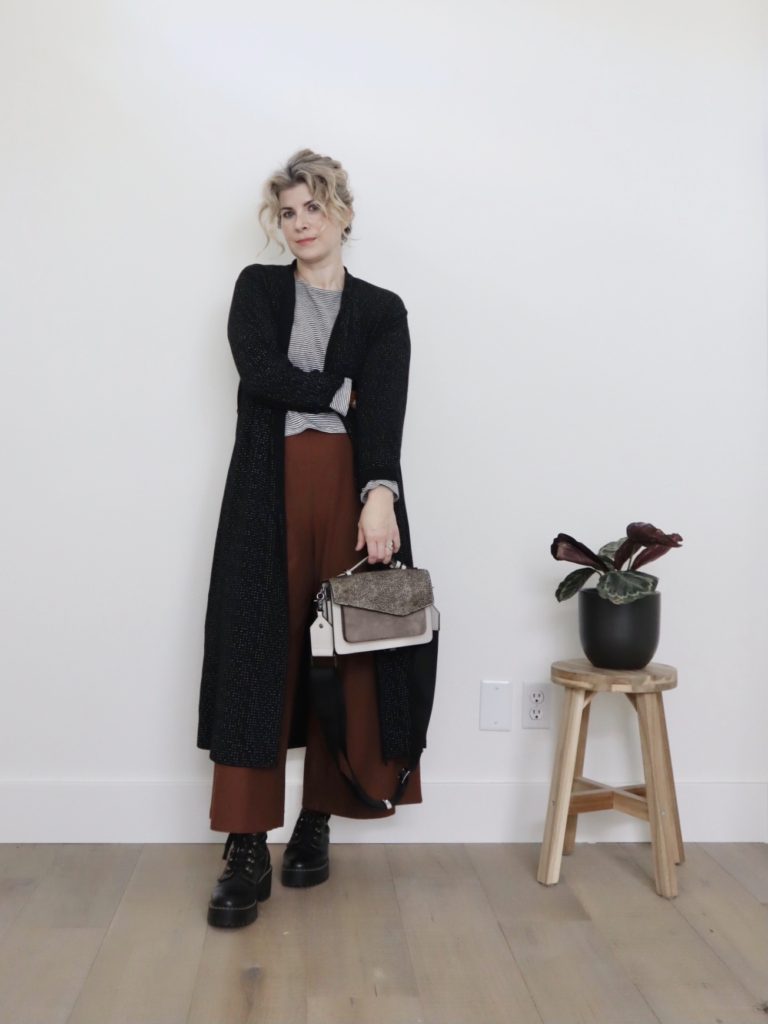 A small white woman is wearing a casual chic outfit the includes a long black duster over a black & white striped top with brown wide leg pants. She is holding a grey handbag and wearing black combat boots.