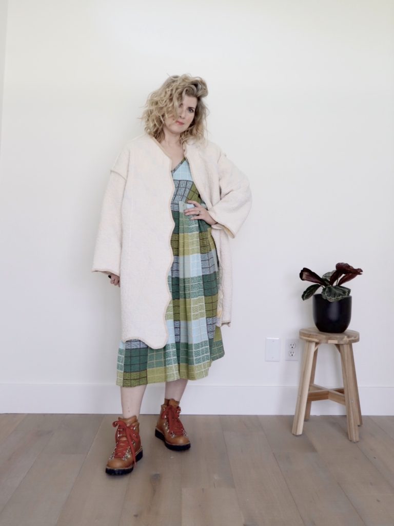 A small white woman is wearing a cream colored quilted coat with tan edging over a colorful midi length dress. The dress is blue green and brown plaid. She is wearing tan boots with red strings.