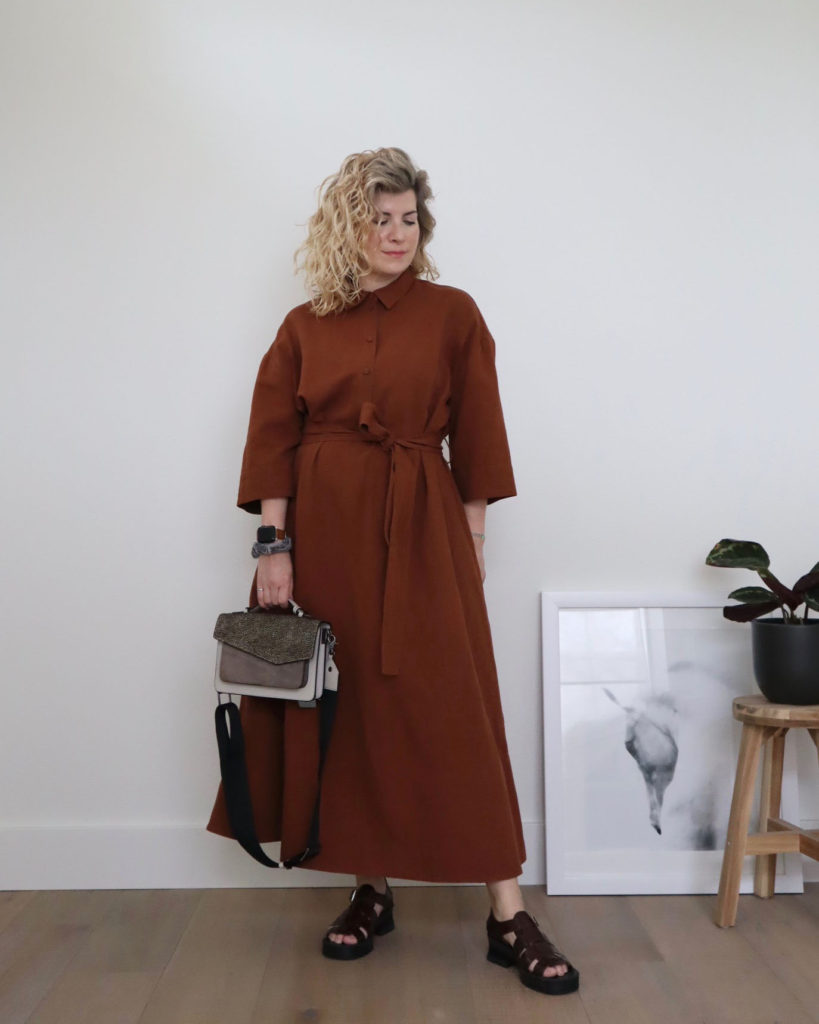 A while woman with blond wavy hair is standing in front of a white wall and wearing a full length brown dress that is belted at the waist, the first of her casual outfits this week. She is wearing brown sandals and holding a grey handbag.