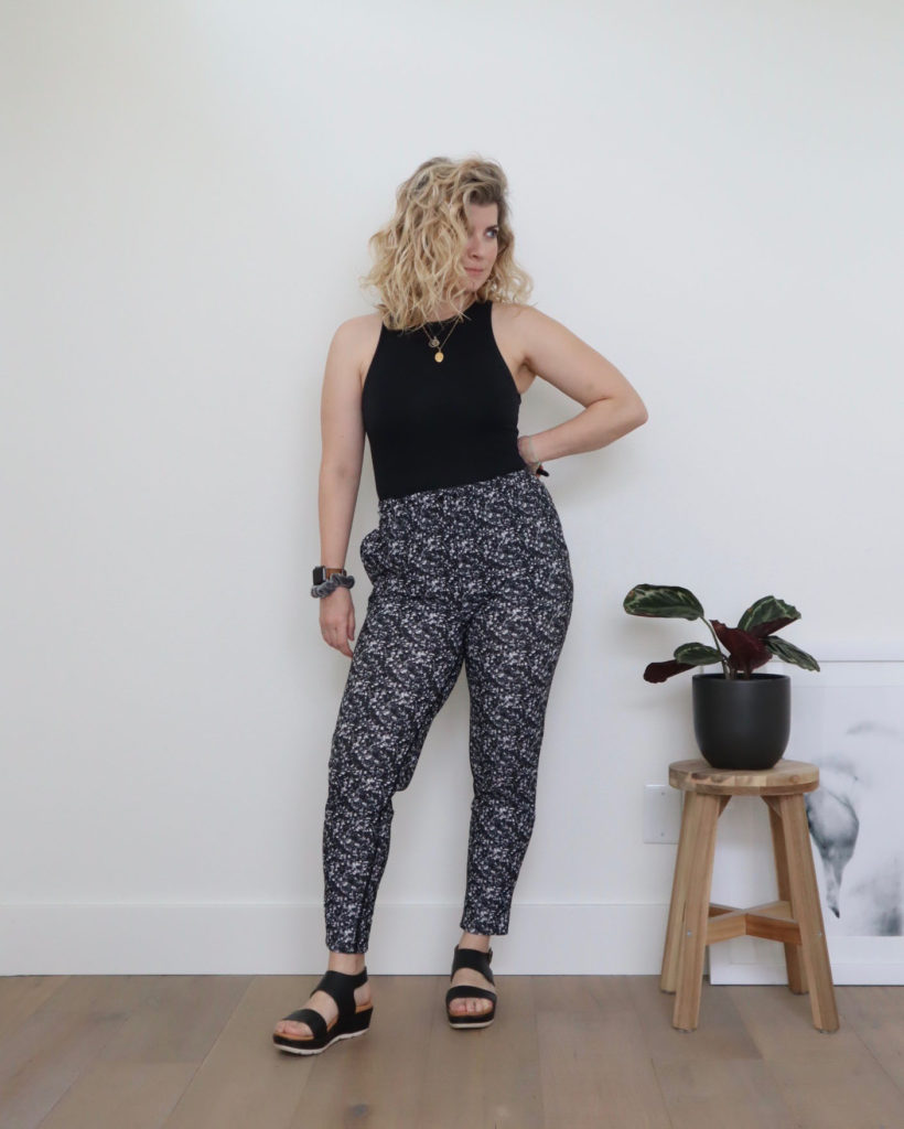 A while woman with blond wavy hair is standing in front of a white wall and wearing a black sleeveless bodysuit with a pair of black and white printed pants, the second of her casual outfits this week. she is wearing black sandals and she has her left hand on her hip.