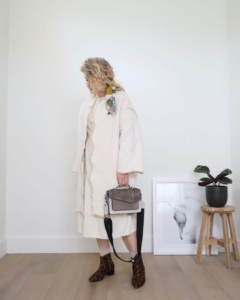 A white woman with blond wavy hair  is wearing a white coat over a white dress and holding a grey handbag, the fifth of her casual outfits this week. She has a cream, mustard, grey and orange print scarf in her hair. She is wearing skin toned socks and black and brown leopard print boots.