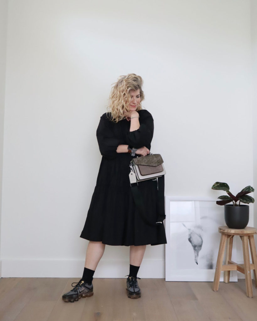 A while woman with blond wavy hair is standing in front of a white wall and wearing a long sleeve black tiered midi dress with black socks and black and gold trainers, the fourth of her casual outfits this week. She has an arm across her body and is holding a grey handbag which the other elbow is resting on her folder arm and her head is resting on her elevated hand.
