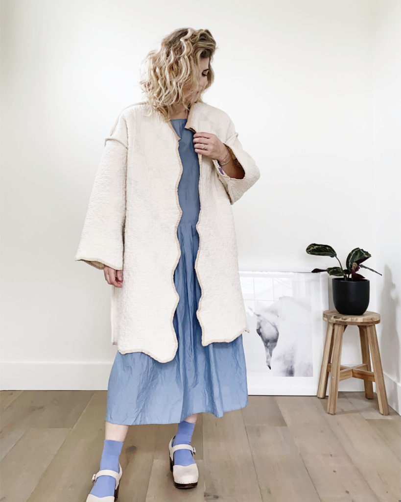 A white woman with blond wavy hair is wearing the coat with the neutral side on the outside over a blue dress with matching blue socks and cream colored clogs.