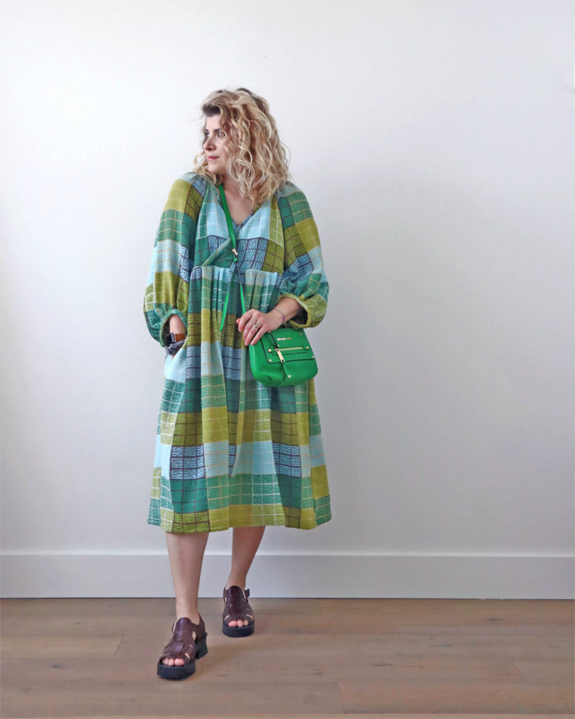 Spring style is color! A white woman with blond wavy hair is standing in front of a light colored wall and wearing a colorful midi dress of green, blue and brown. She had a green crossbody bag across her chest and is wearing brown chunky sandals with thick straps.