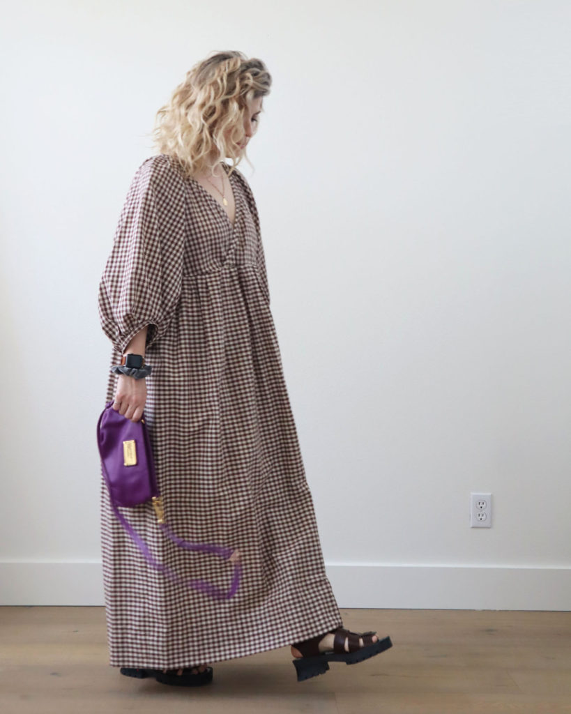 Spring style is a loose, flowy dress. A white woman with blond wavy hair is in motion in front of a light colored wall. She is wearing a long dress that is brown and white checkered with brown chunky sandals and is holding a purple crossbody bag in her right hand.