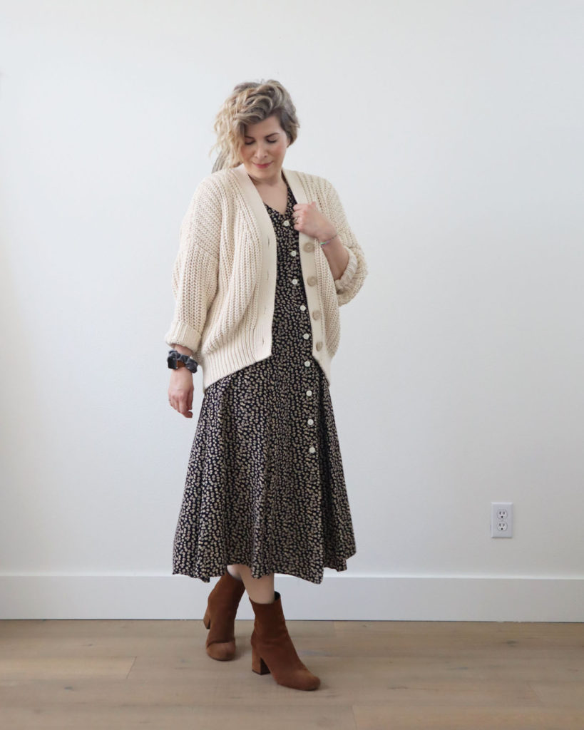 Spring style is cozy. A white woman with blond wavy hair is standing in front of a light colored wall and wearing a cream colored chunky cardigan open over a navy floral midi dress with cream buttons up the front. She is wearing brown heeled boots.