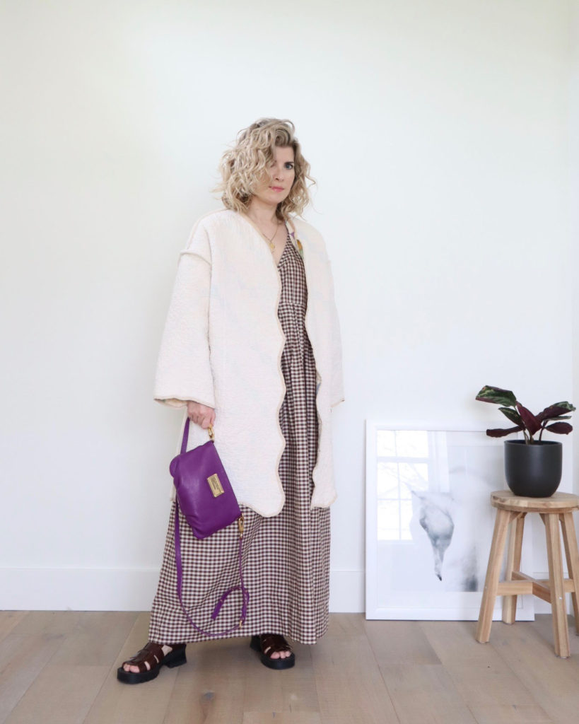 Spring outfits, week 4, look 1 - a small white woman is wearing a knee length white quilted coat over a long brown and white gingham dress and brown chunky sandals. the it holding a small purple handbag in her right hand but the strap. She is turned slightly sideways and looking at the camera.