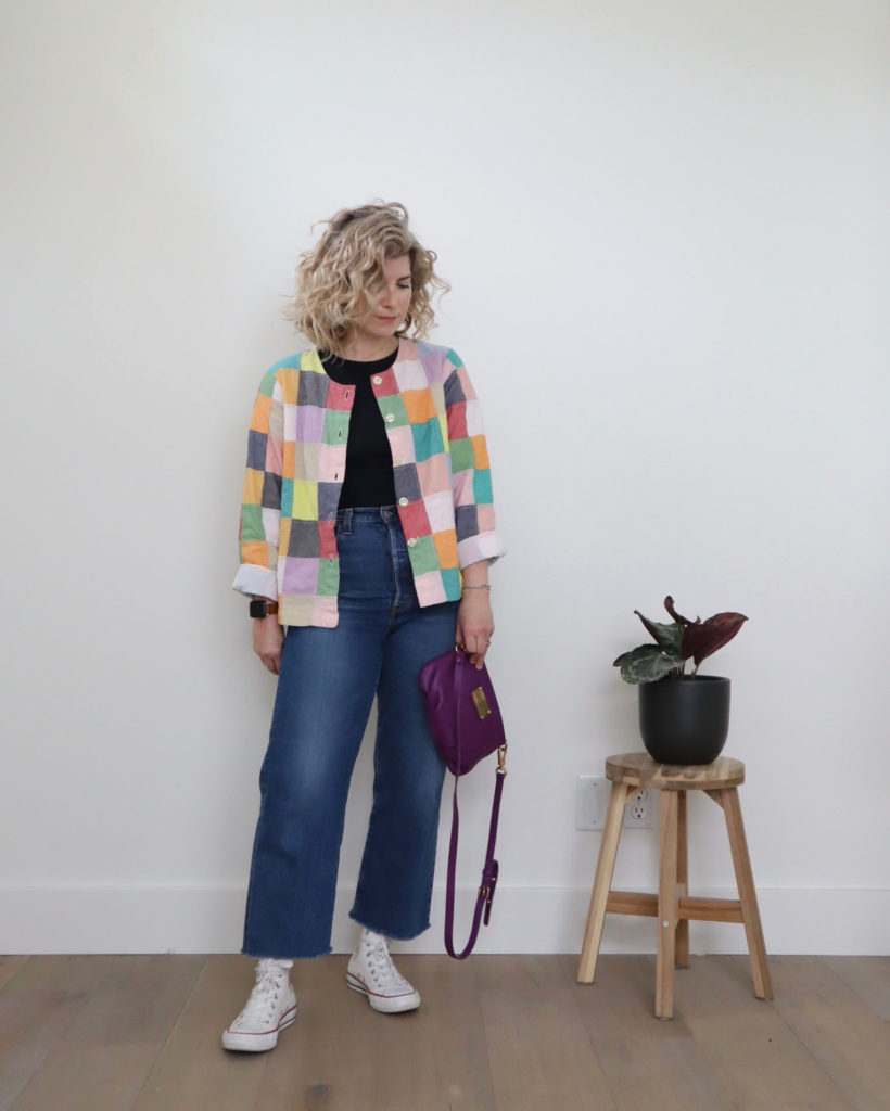 Spring outfits, week 4, look 4 - a small white woman is wearing a patchwork jacket made up of pastel colors. The jacket is open over a black bodysuit and medium wash blue jeans. She is wearing white converse high top sneakers and holding a purple handbag.