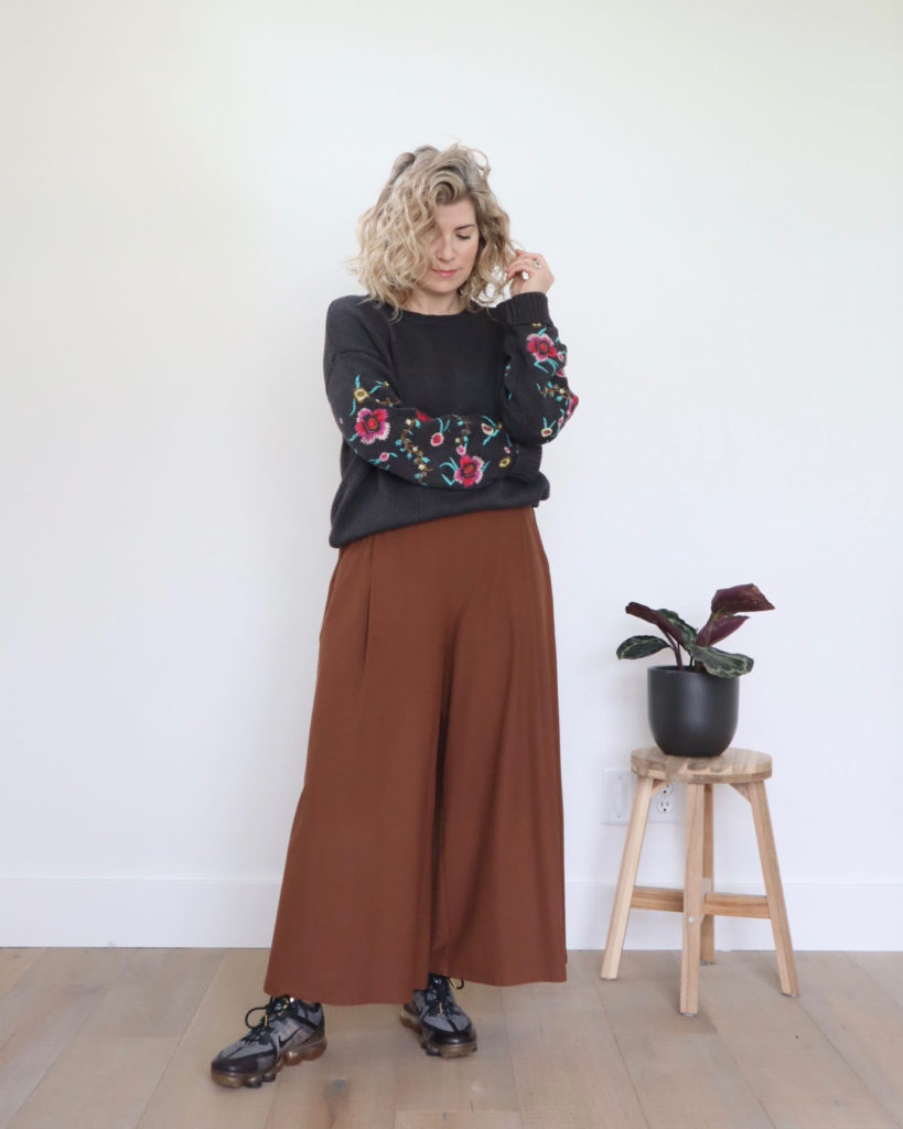 Spring outfits, week 4, look 5 - a small white woman is wearing a a grey long sleeved sweater with floral embroidery down the sleeves. She is wearing it with brown wore legged pants and black, grey and gold trainers.