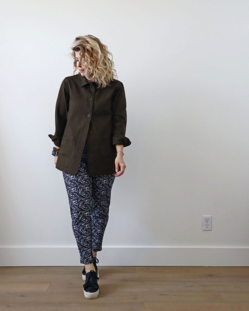 Spring style is versatile. A white woman with blond wavy hair is standing in front of a light colored wall and wearing an olive green coat buttoned all the way to the to with the bottom buttons left undone. She is wearing it with a pair of black and white pattered pants and black platform sneakers with a thick white sole.