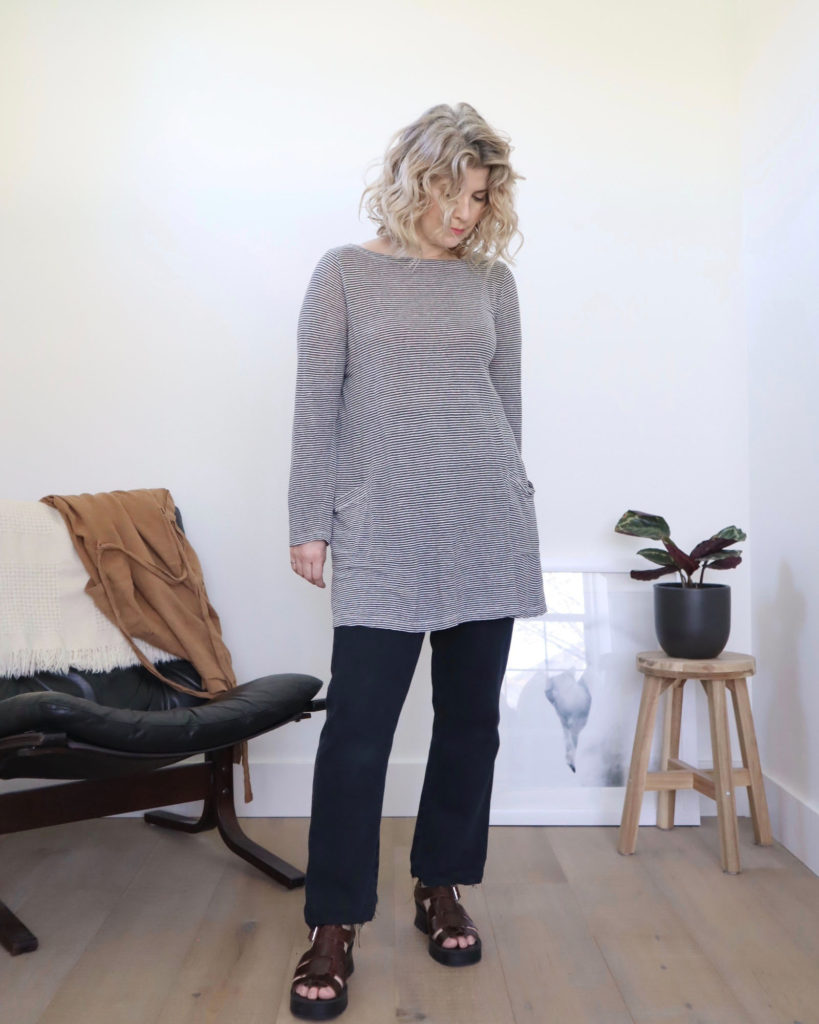A white woman with short blonde wavy hair is looking down towards the floor. She is wearing a long sleeved black and white tunic over black jeans with brown strappy platform sandals. There is a black chair behind her to the left and a prayer plant on a wooden stool behind her to the right. She is using this top as a tutorial on how to crop a top.
