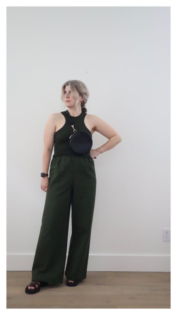 A small white woman with blonde hair is wearing a simple outfit of an olive green tank top with olive green wide leg pants. She has a small circle bag across her body and is wearing brown scrappy sandals with a thick black sole.