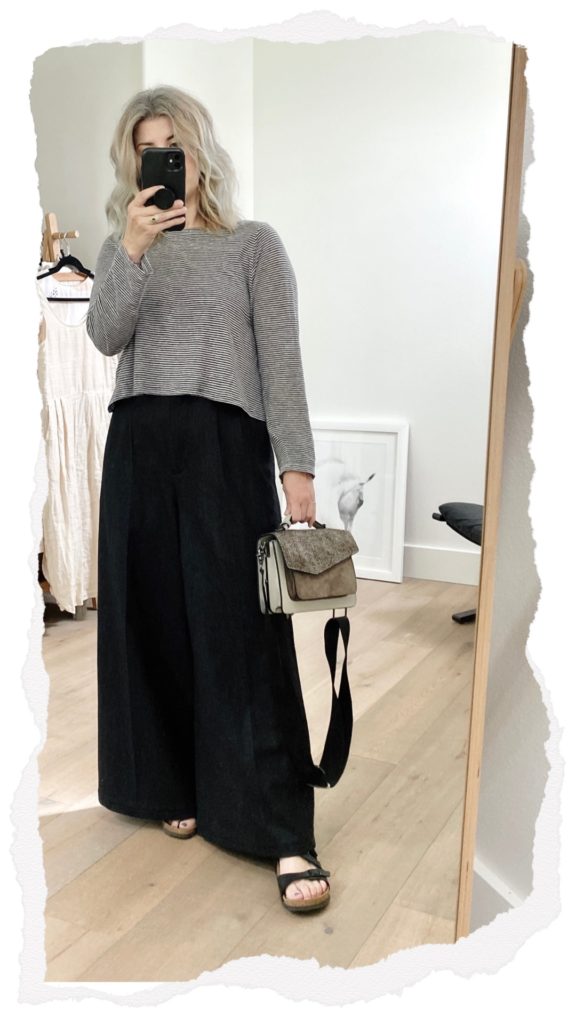 A mirror selfie of a small white woman  who is wearing a waist length striped long sleeve top with a pair of wide legged dark grey trousers. she is wearing black sandals and holding a grey handbag.