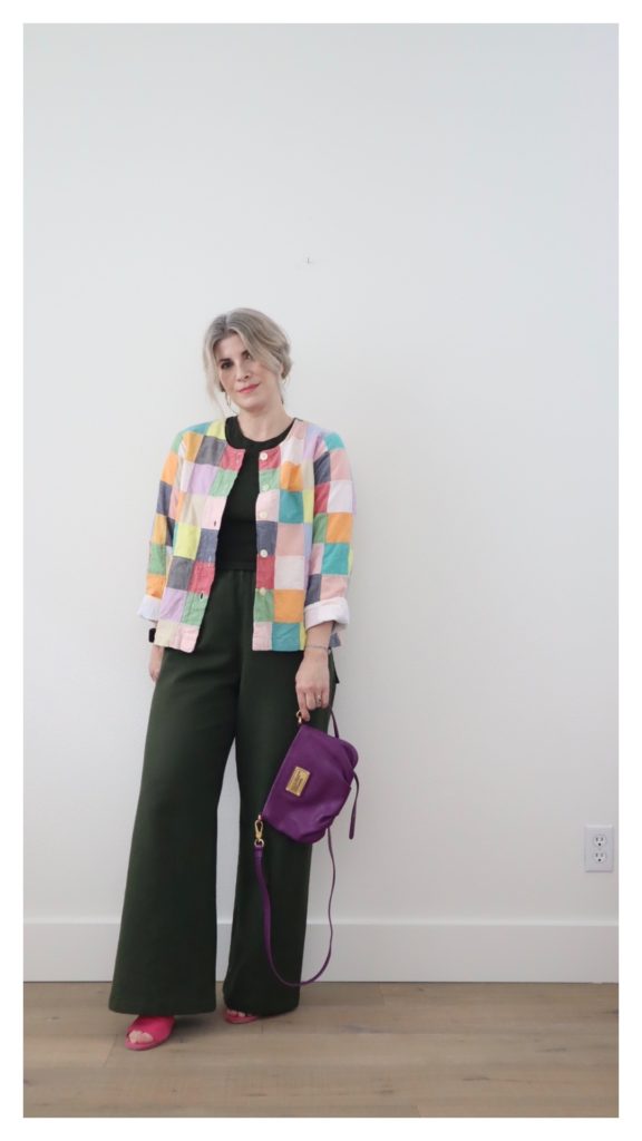 A small white woman with blonde hair is wearing a simple outfit of an olive green tank top with olive green wide leg pants. She is wearing a patchwork jacket of pastel colors and hot pink sandals. She is holding a purple handbag in the left hand.