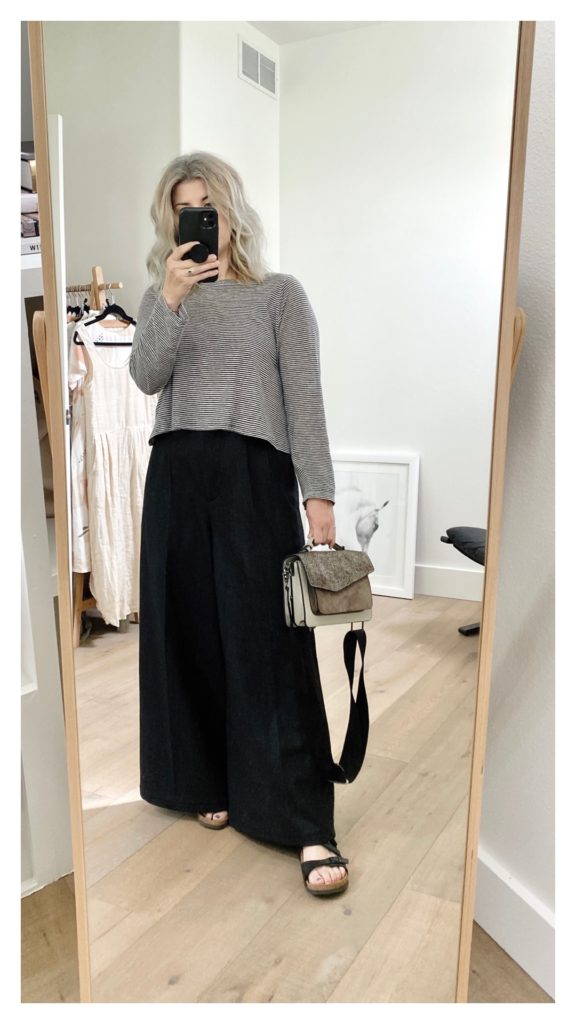 A mirror selfie of a small white woman with shoulder length blond hair is wearing a waist length black and white striped long sleeve top with a pair of grey wide leg pants and black sandals. She is holding a grey handbag in her right hand.